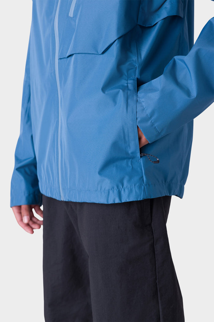 A person wears the 686 Men's Hydra™ 2.5L All-Weather Jacket—blue, with their left hand in the pocket—and black pants. Only the lower half of the jacket and upper legs show against a white background.