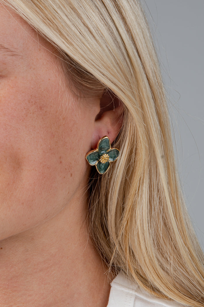 A close-up of a blonde woman's ear wearing a gold four-petal flower stud earring with green enamel details.