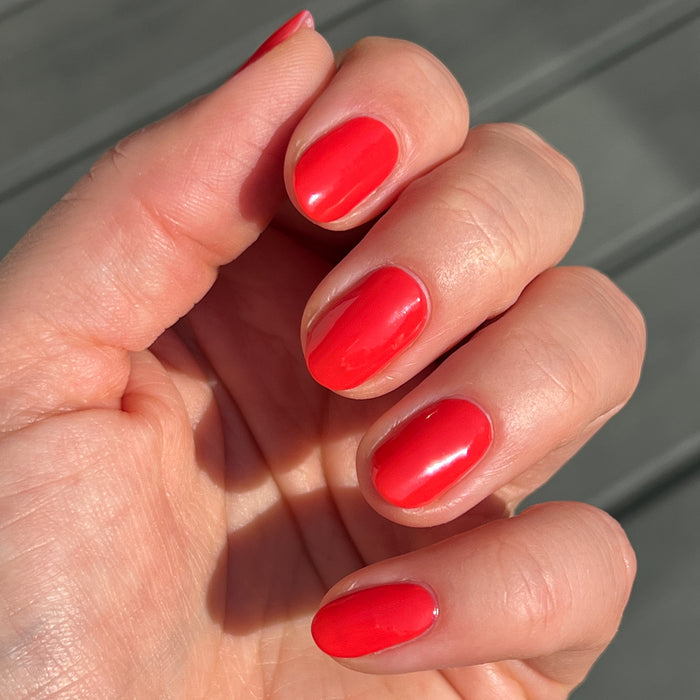 Hand with nails polished in glossy orangey red Picante polish shown in natural sunlight for manicure display