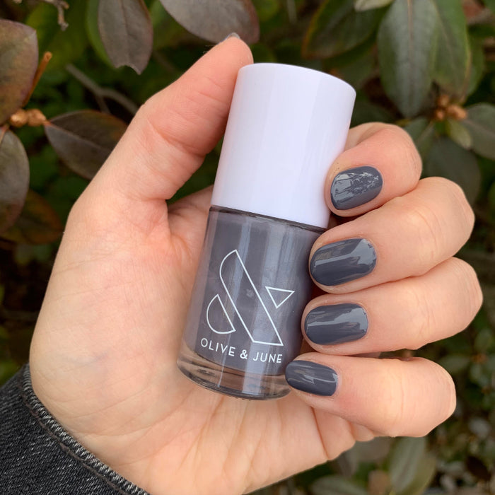 Hand holding Olive and June 100% CHANCE polish bottle with nails painted in deep gunmetal blue for a smooth mani