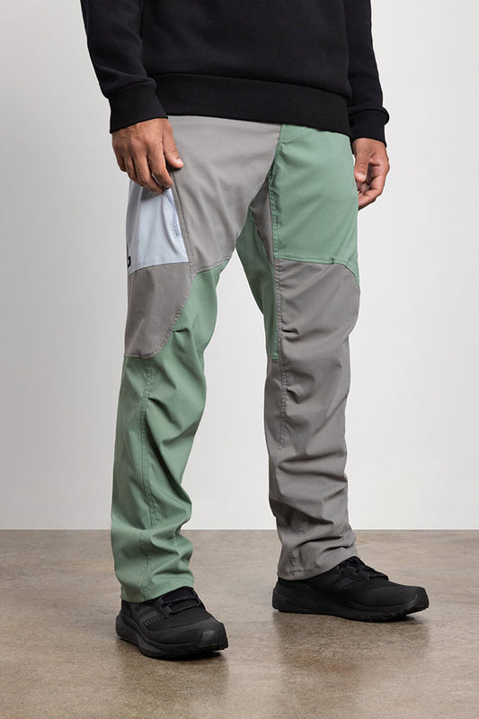 A person stands on a concrete floor against a light background, wearing 686 Mens Anything Cargo Pant - Relaxed Fit by 686 in green and gray color-block with 4-way stretch, paired with a black sweatshirt and black shoes.