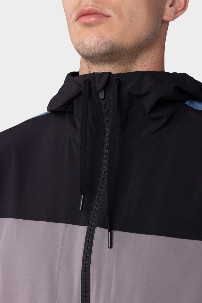 A close-up of a person wearing the 686 Mens ATP Stretch Wind Jacket, featuring black and gray color-blocking and a hood with black drawstrings. The persons face is partially visible from the nose down.