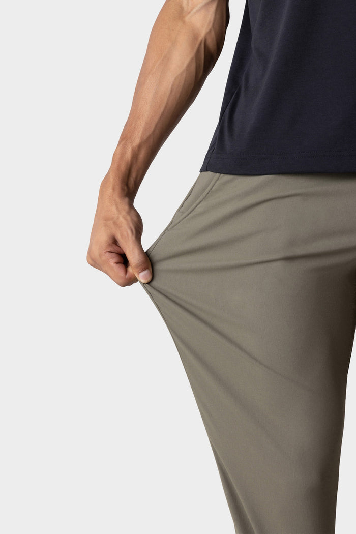 A person in 686 Mens Everywhere Merino-Lined Pant - Slim Fit stretches the olive green fabric with one hand, demonstrating its flexibility and warmth against a plain white background.