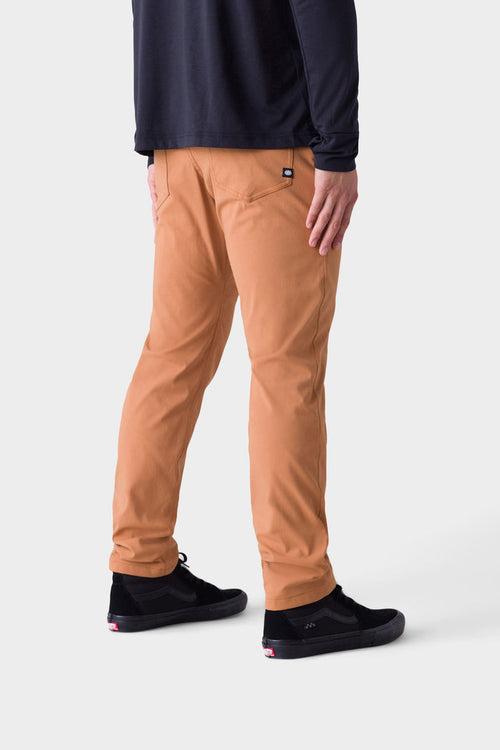 A person faces away, wearing 686 Mens Everywhere Pant - Slim Fit in tan, a long-sleeved black shirt, and black sneakers with white soles, set against a plain white background.