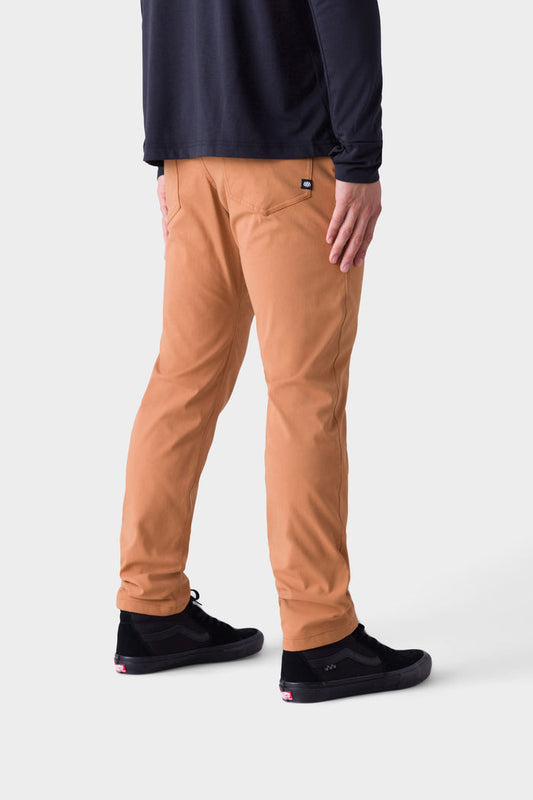 A person faces away, wearing 686 Mens Everywhere Pant - Slim Fit in tan, a long-sleeved black shirt, and black sneakers with white soles, set against a plain white background.