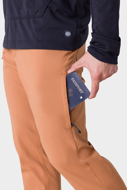 A person wearing the 686 Mens Everywhere Pant - Slim Fit in tan places a navy passport into the pants zippered side pocket. The background is plain white.