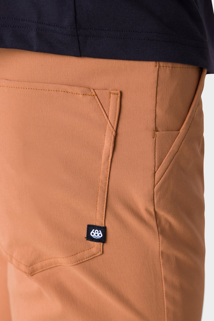 Close-up of a person wearing 686 Mens Everywhere Pant - Slim Fit in tan, featuring a visible front pocket and a small black tag with white 686 logo, paired with a dark-colored shirt.