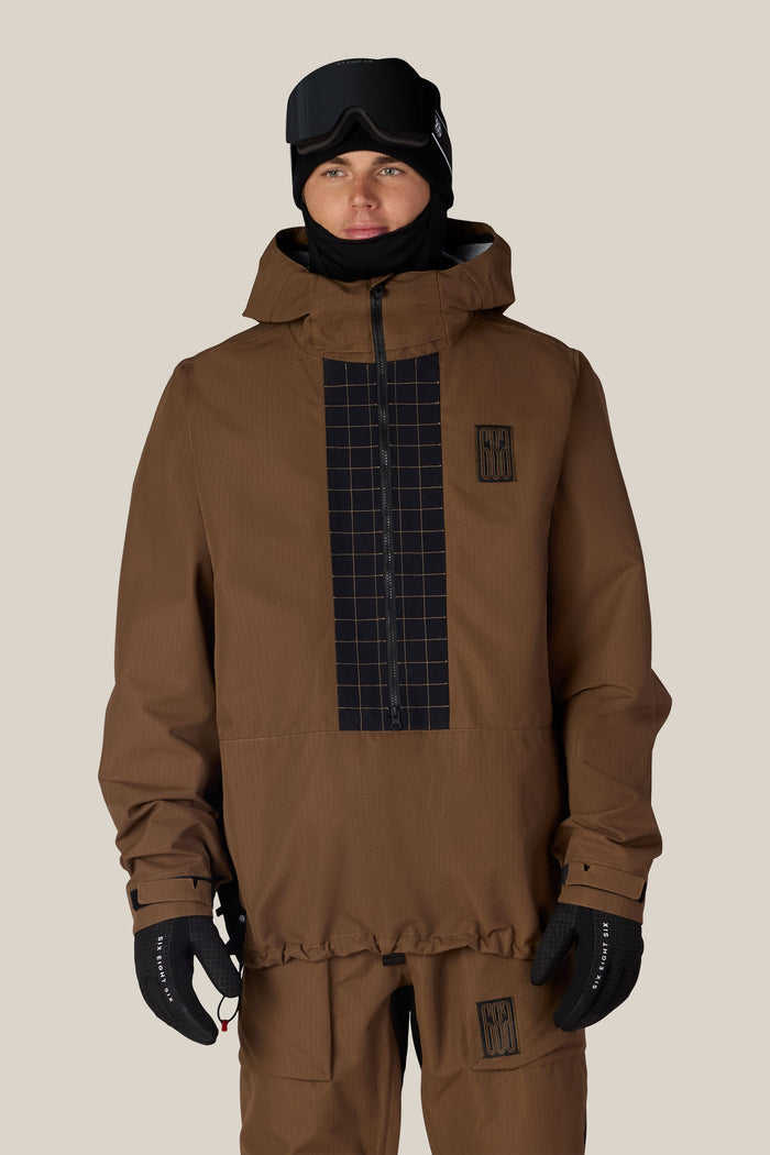 A person wearing the 686 Men’s Ghost™ 2.5L Anorak by 686, featuring a brown color with black grid patterns, paired with matching pants, black gloves, and black ski goggles against a light beige background.