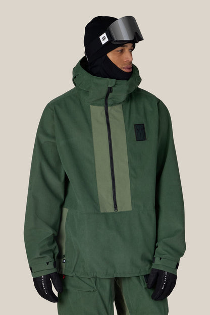 A person wearing the 686 Men’s Ghost™ 2.5L Anorak by 686 in green, paired with matching pants, black gloves, a black beanie, and ski goggles, stands against a plain beige background.