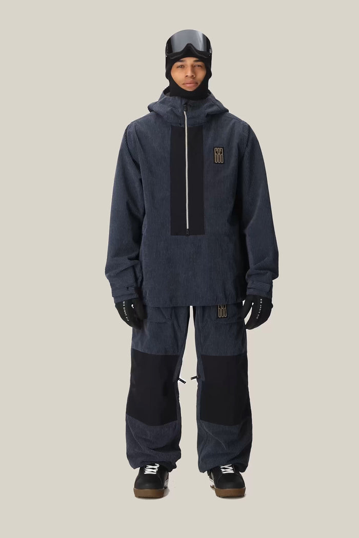A person wearing the 686 Men’s Ghost™ 2.5L Anorak by 686 in dark blue, paired with matching pants featuring black patches, along with gloves, a helmet, and goggles, stands against a light beige background.