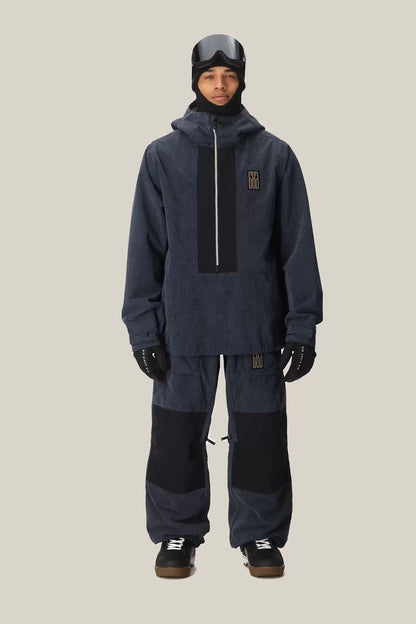 A person wearing the 686 Men’s Ghost™ 2.5L Anorak by 686 in dark blue, paired with matching pants featuring black patches, along with gloves, a helmet, and goggles, stands against a light beige background.