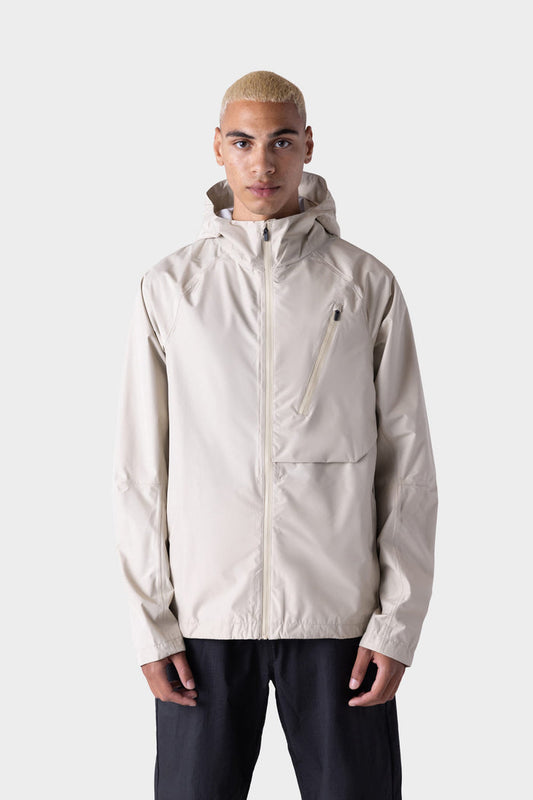A person with short, bleached hair models the 686 Men's Hydra™ 2.5L All-Weather Jacket in light beige, featuring a chest zipper pocket, paired with black pants against a plain light gray background.
