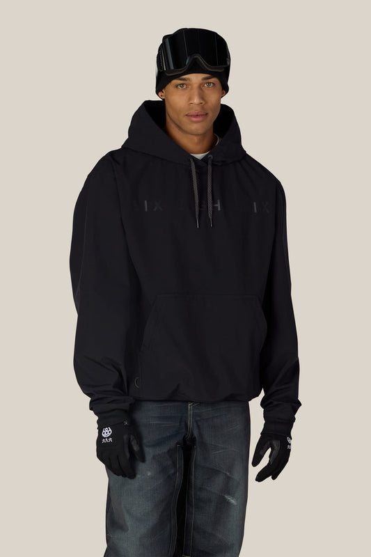 A man wearing the 686 Men’s Waterproof Team Hoody from 686, paired with black gloves, dark jeans, and ski goggles on his forehead, stands against a plain light background and looks at the camera with a neutral expression.