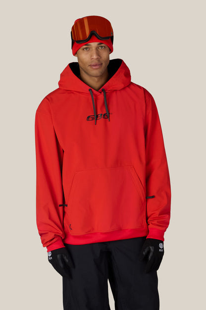 A person wearing the 686 Men’s Waterproof Team Hoody in bright red, paired with a red beanie, orange ski goggles, black gloves, and black pants stands facing forward against a plain light background.