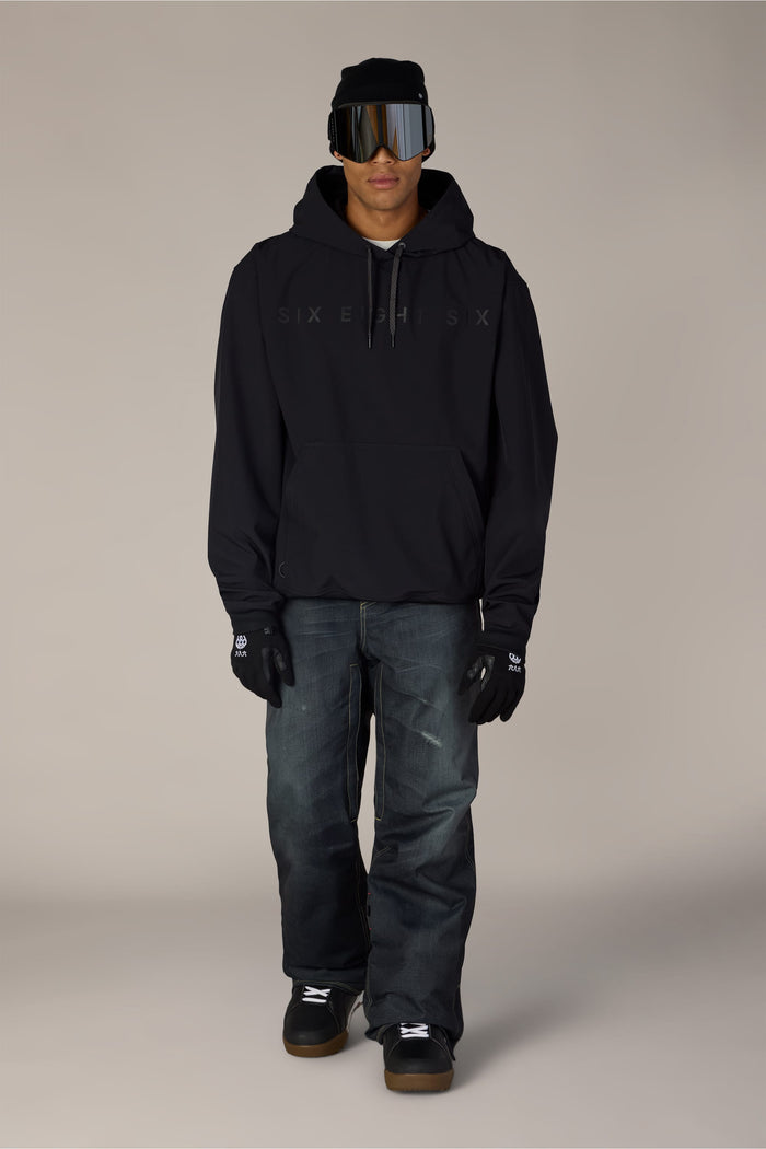 A person is wearing the 686 Men’s Waterproof Team Hoody by 686, along with a black beanie, reflective ski goggles, black gloves, dark jeans, and black sneakers, standing against a plain light background.