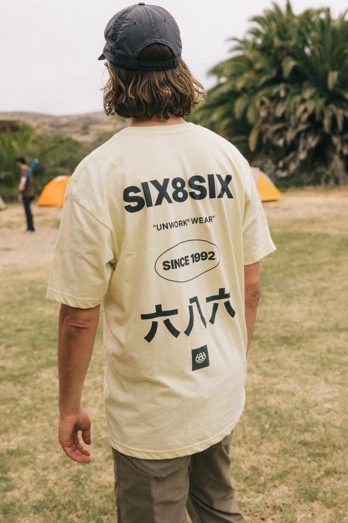 A person with wavy hair in a cap and a light-colored 686 Unwork Wear Authentic S/S Tee, featuring ‚ÄúSIX8SIX,‚Äù ‚ÄúUNWORK WEAR,‚Äù ‚ÄúSINCE 1992,‚Äù and Chinese characters on the back, stands on grass near orange tents and palm trees.