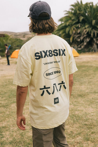 A person with wavy hair in a cap and a light-colored 686 Unwork Wear Authentic S/S Tee, featuring ‚ÄúSIX8SIX,‚Äù ‚ÄúUNWORK WEAR,‚Äù ‚ÄúSINCE 1992,‚Äù and Chinese characters on the back, stands on grass near orange tents and palm trees.
