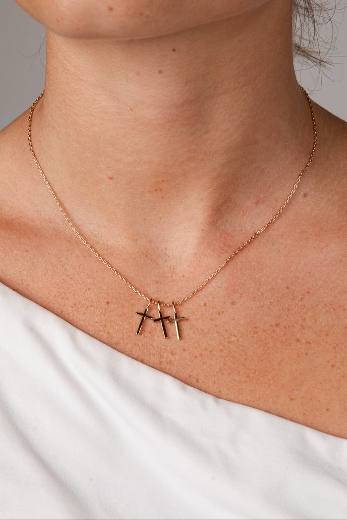 A close-up of a model's neck and upper chest wearing a delicate rose gold chain necklace with three small vertical cross pendants.
