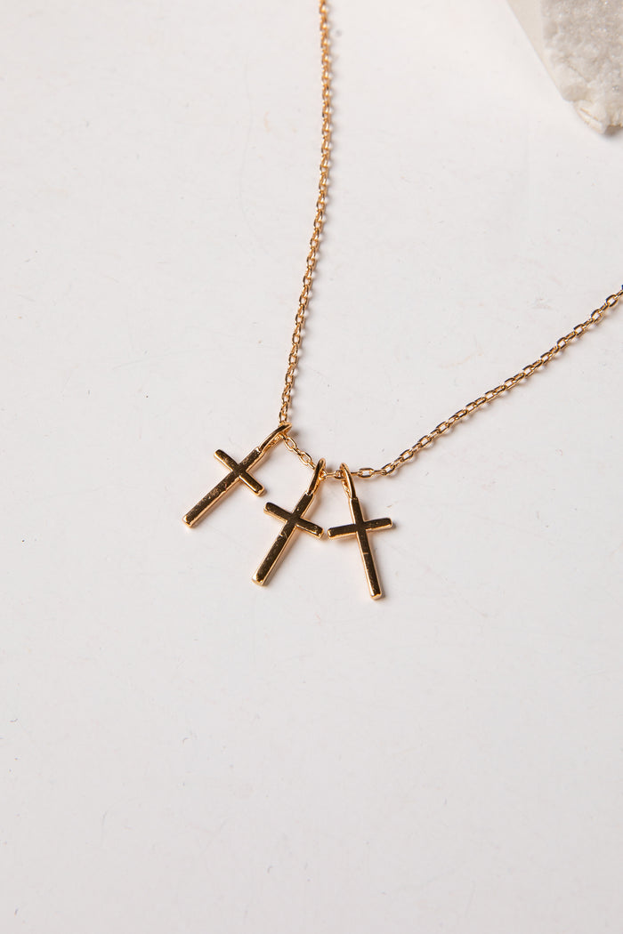 A gold chain necklace with three small cross pendants in a gold-tone finish, displayed on a light off-white surface.