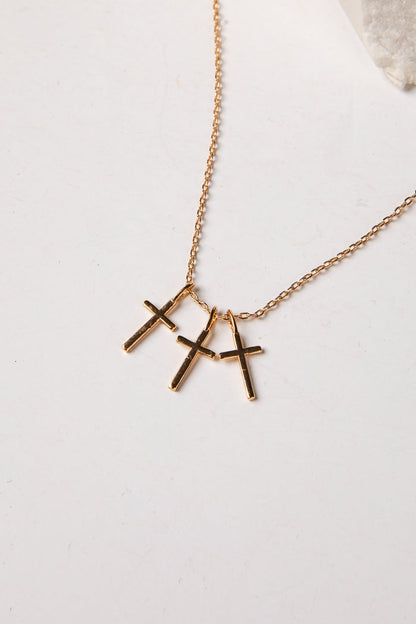 A gold chain necklace with three small cross pendants in a gold-tone finish, displayed on a light off-white surface.