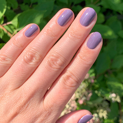 Hand with glossy muted eggplant nails showing Art Class polish manicure outdoors with green leaf background