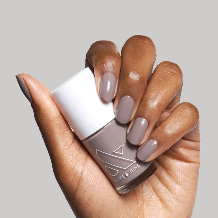 Hand with nails painted in warm grey AW shade holding Olive & June nail polish bottle against light grey background