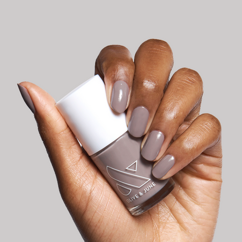 Hand with nails painted in warm grey AW shade holding Olive & June nail polish bottle against light grey background