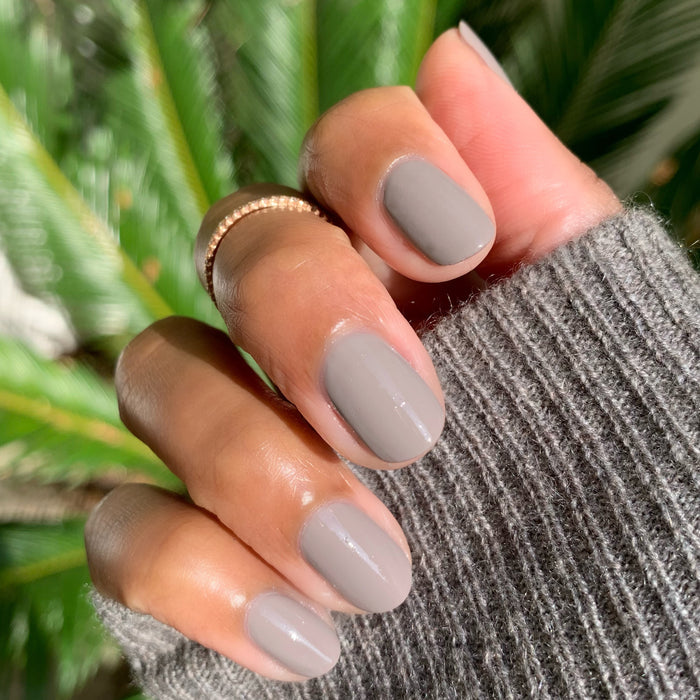Fingers with nails painted in AW warm grey polish showing a smooth, glossy mani against a green plant background