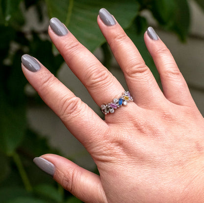 Hand with nails painted in AW warm toned grey showing a smooth manicure with a colorful gemstone ring against a leafy background