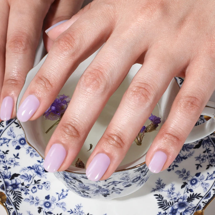 A hand with dreamy Cloud Nine by LONDONTOWN periwinkle nails rests on a floral teacup and saucer filled with water and small purple flowers.