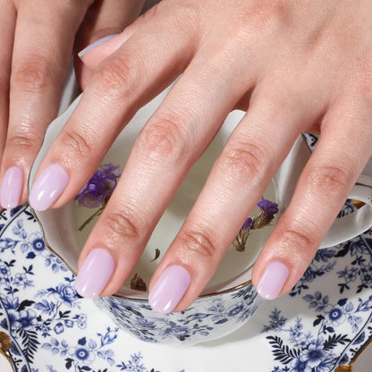 A hand with dreamy Cloud Nine by LONDONTOWN periwinkle nails rests on a floral teacup and saucer filled with water and small purple flowers.