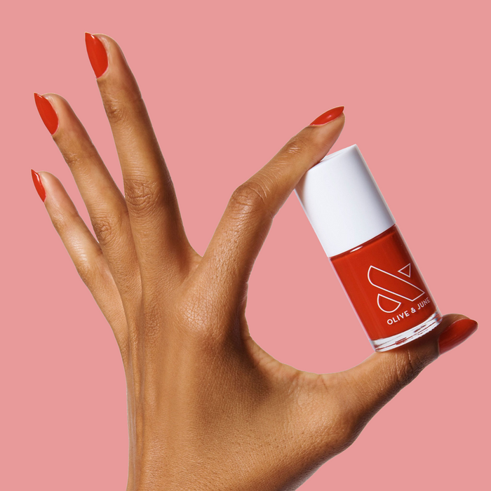 Hand with nails painted bright cherry red holding Olive and June CV polish bottle against pink background