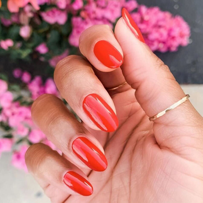Hand with nails painted in glossy bright cherry red shade CV, shown outdoors with pink flowers in the background