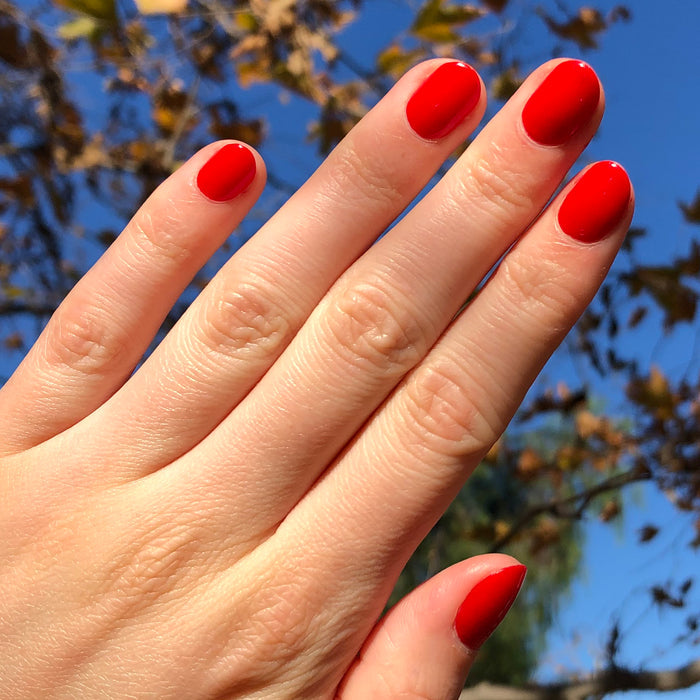 Hand with nails painted in bright cherry red CV polish by Olive and June shown outdoors against autumn foliage backdrop