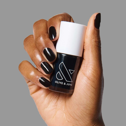 Hand with nails painted in deep black holding a bottle of E+M nail polish by Olive and June against gray background