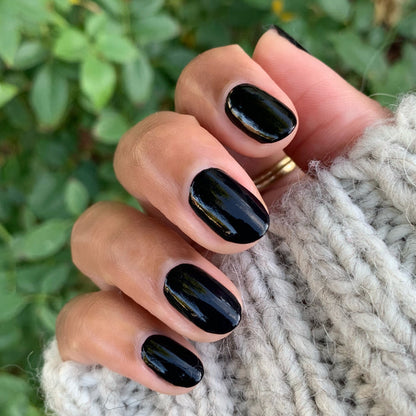 Hand with nails painted in glossy deep black E+M polish by Olive and June against natural green background