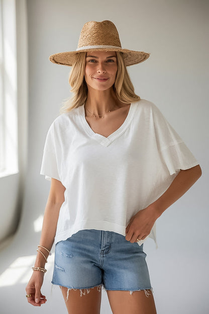 A woman wearing a white short-sleeve V-neck top, light blue denim shorts, and a straw hat, standing in a brightly lit indoor setting.