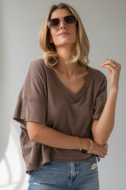 A woman wearing a brown short-sleeve V-neck t-shirt and blue jeans, styled with sunglasses, bracelets, and a necklace, posing against a neutral gray background.