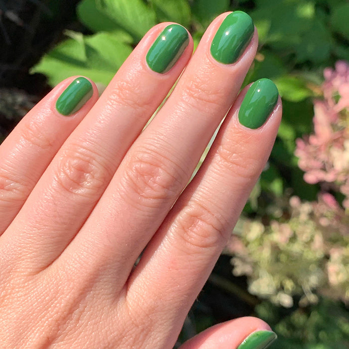 Hand with a fresh manicure featuring Geometry polish in vibrant hunter green against a natural outdoor background