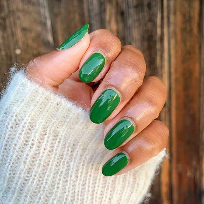 Hand with a glossy Geometry polish manicure in deep hunter green against a cozy off-white sweater and wooden background