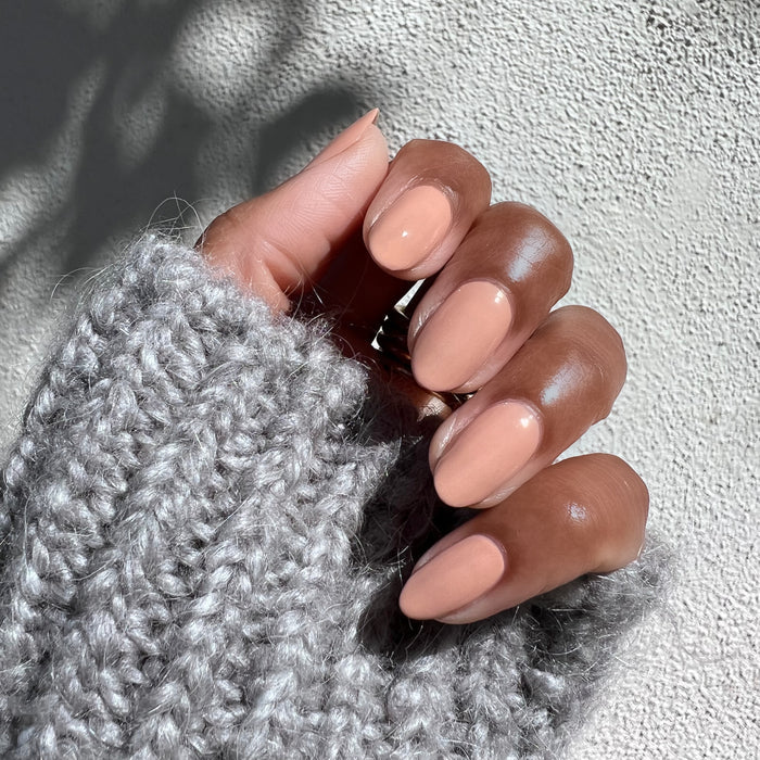Hand with nails painted in Howdy neutral beige polish showing a smooth glossy manicure against textured gray knit background