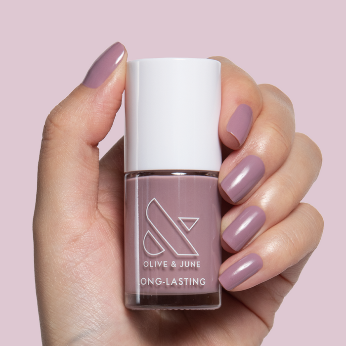 Hand with midtone mauve nails holding Olive and June I Mani in Meetings long-lasting polish bottle against light pink background