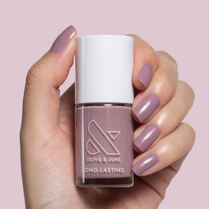 Hand with midtone mauve nails holding Olive and June I Mani in Meetings long-lasting polish bottle against light pink background