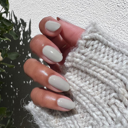 Hand with nails painted in Koala soft grey shade resting on cozy knit fabric in natural sunlight for a polished mani