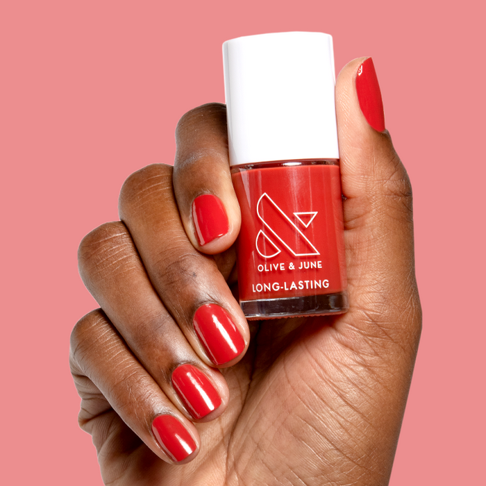 Hand holding Olive and June Let’s Circle Back long-lasting gel polish bottle with matching bright orange-red mani on pink background
