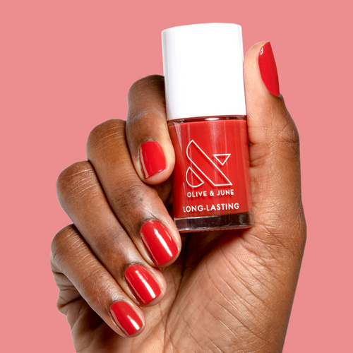 Hand holding Olive and June Let’s Circle Back long-lasting gel polish bottle with matching bright orange-red mani on pink background