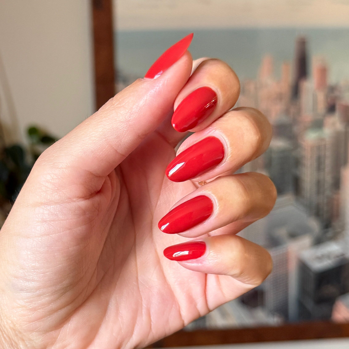 Hand with glossy Let’s Circle Back bright orange-red gel polish manicure shown against blurred cityscape background