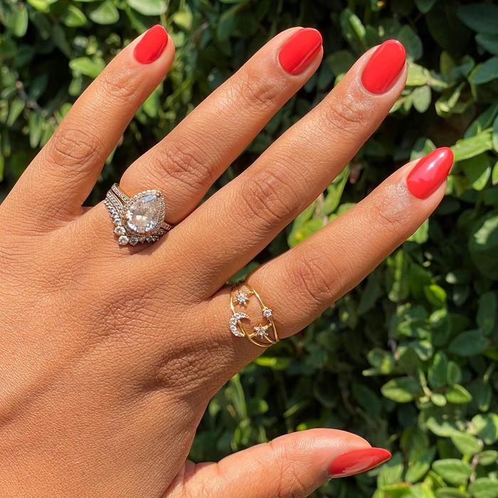 Hand with glossy bright orange-red Let’s Circle Back gel polish manicure shown outdoors against green leaves background