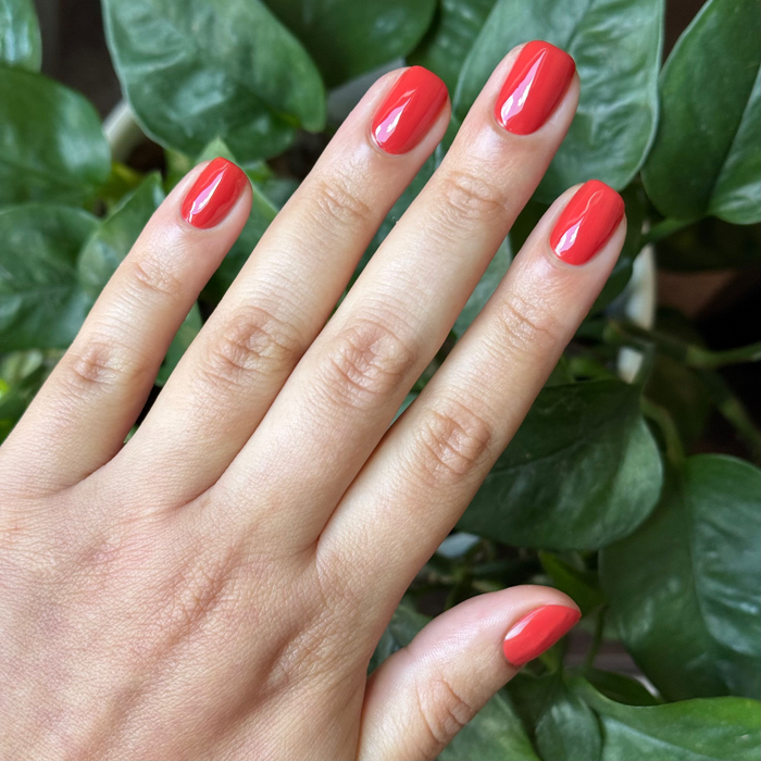 Hand with glossy bright orange-red Let’s Circle Back gel polish manicure shown against green leafy background
