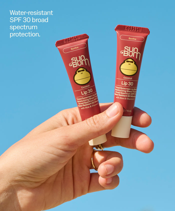 Hand holding two Sun Bum Tinted Lip Duos tubes in Bonfire shade against blue sky background. Water-resistant
SPF 30 broad spectrum protection.